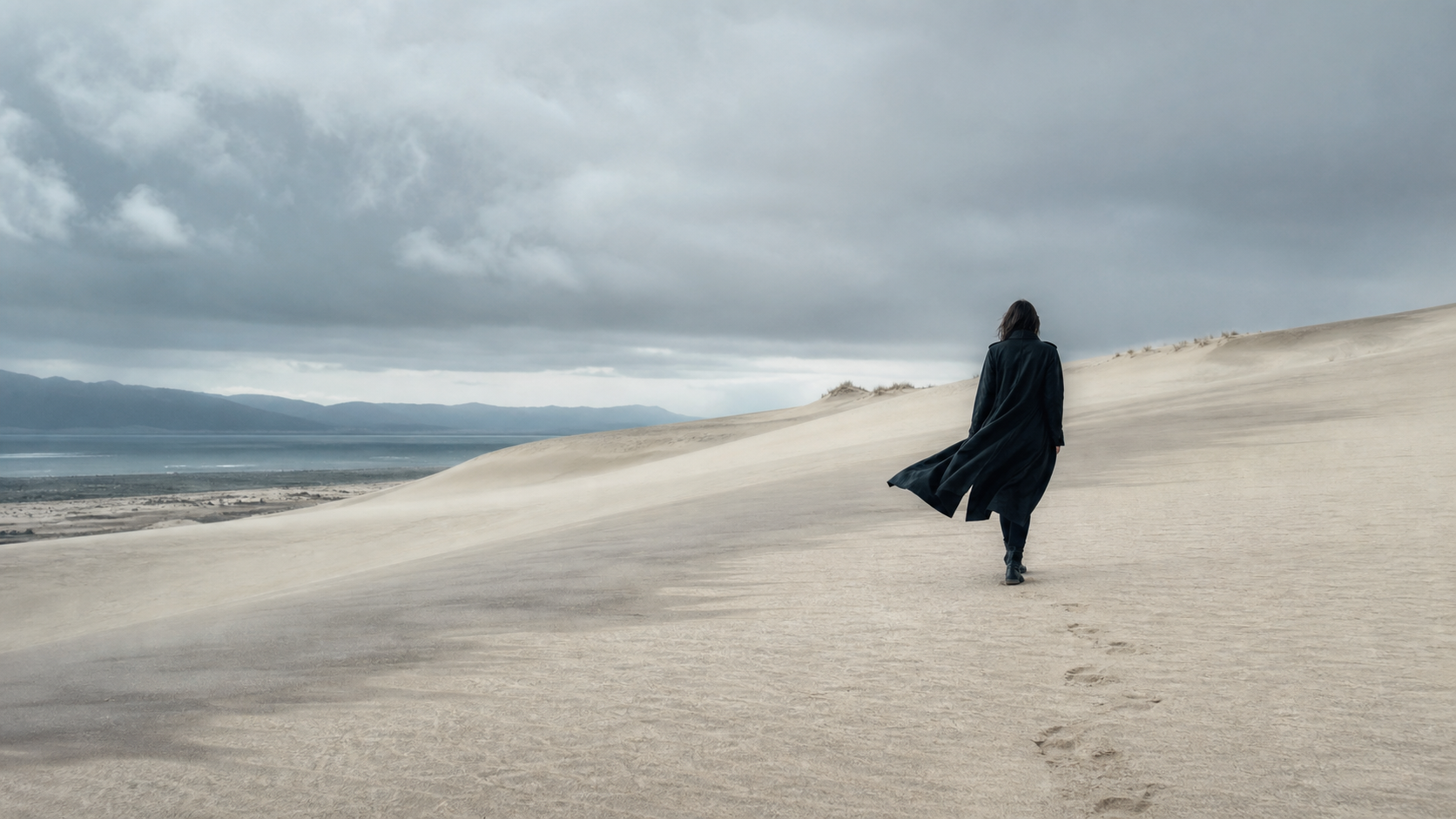 Person in dunklem Mantel in einer weiten Dünenlandschaft